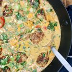 A skillet filled with creamy coconut curry with golden-brown pork meatballs, vibrant bell peppers, sweet potatoes, and fresh spinach. The dish is garnished with chopped cilantro, green onions, and peanuts, with a silver serving spoon resting in the pan.