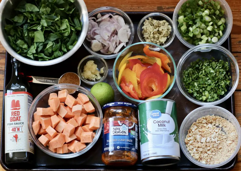 A flat lay of ingredients for the curry, including sweet potatoes, bell peppers, spinach, Thai red curry paste, coconut milk, and fish sauce, all arranged on a baking sheet.