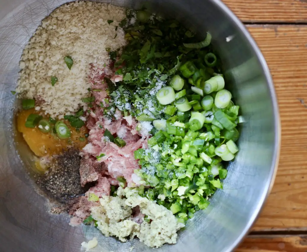 A close-up of a stainless steel mixing bowl filled with ingredients for pork meatballs, including ground pork, panko breadcrumbs, egg, chopped green onions, cilantro, minced garlic, ginger, and seasonings.