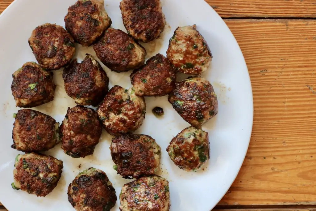 A white plate filled with freshly seared pork meatballs, golden-brown on the outside with flecks of green from fresh herbs. The plate sits on a rustic wooden table.