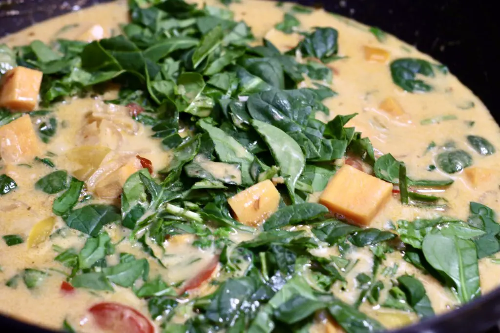 A close-up of coconut curry simmering in a pan, with fresh spinach, sweet potatoes, and bell peppers floating in a rich, creamy sauce. The spinach is just starting to wilt into the curry.