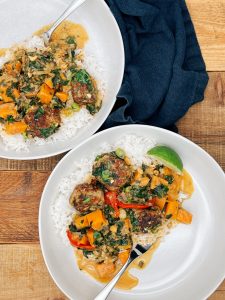Two white bowls filled with coconut curry pork meatballs, served over fluffy rice with chunks of sweet potato, spinach, and red bell pepper. A fresh lime wedge sits on the side, and a spoon rests in one of the bowls. A dark blue napkin adds contrast to the rustic wooden background.